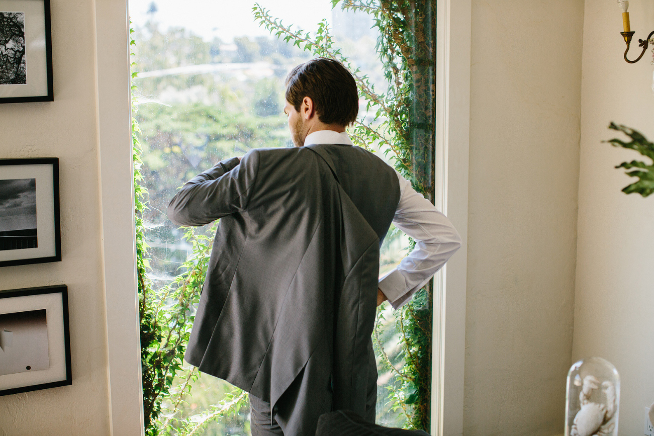 The groom putting on his jacket by a window. 
