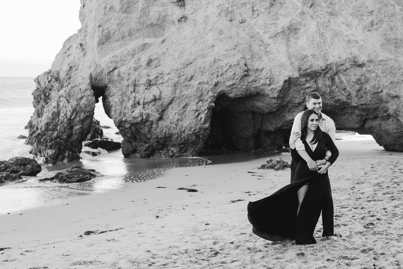 black and white photo with rocks at the beach in the background. His arms wrapped around his bride.