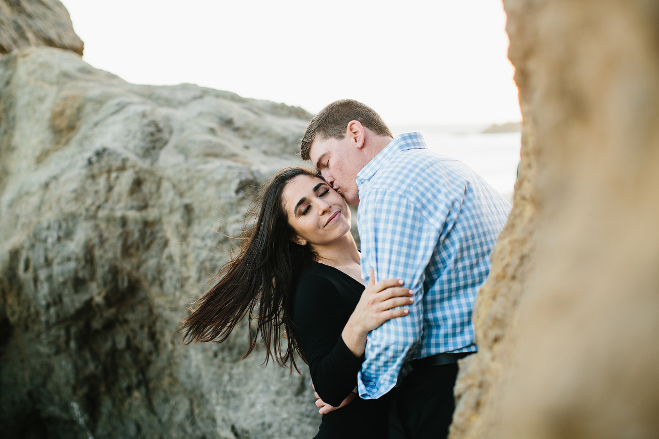 down on the beach groom to be kissing fiances cheek as her hair blows in the wind.