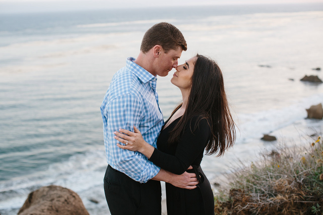 engaged couple cliffside nise to nise faces close and eyes closed with the gorgeous beach in the background
