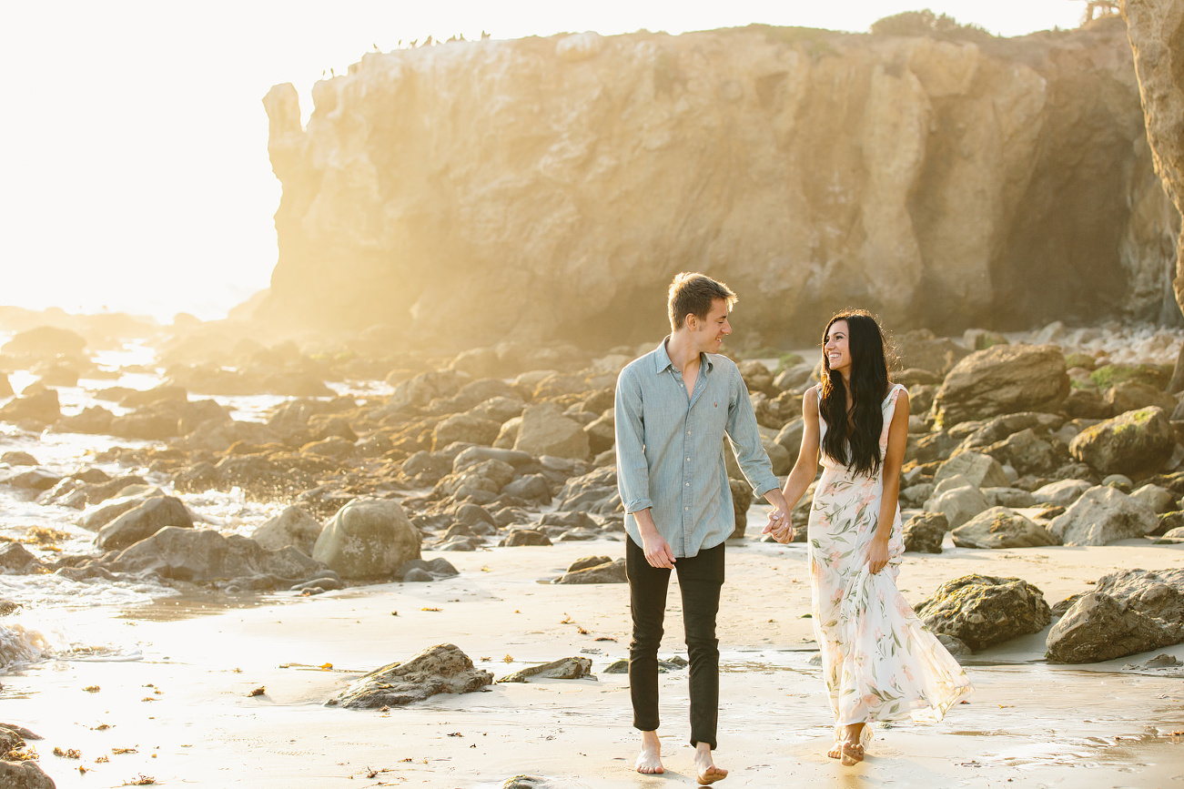 Engaged couple walking on the beach with rocks behind them